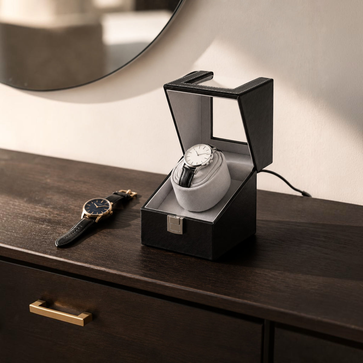 Remontoir montre automatique noir posé sur une commode en bois sombre, couvercle relevé et câble branché