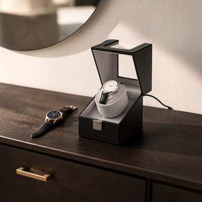Remontoir montre automatique noir posé sur une commode en bois sombre, couvercle relevé et câble branché